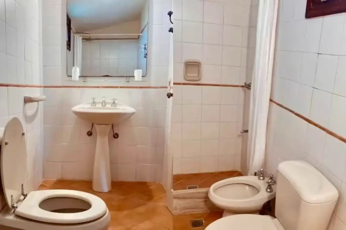 Baño con azulejos blancos, lavabo con pedestal, ducha con cortina y bidé