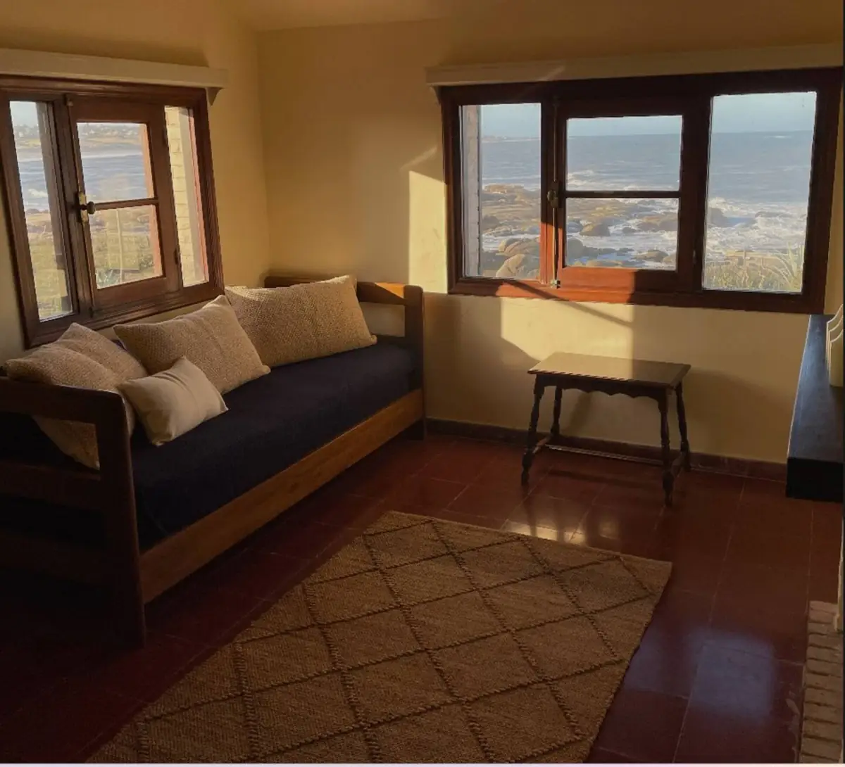 Cama de día con almohadones junto a dos ventanas con vista al océano y las rocas